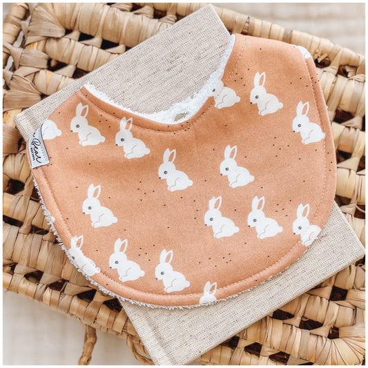 Handmade baby bib with a terracotta background and repeating white bunny design. Bib is backed with beige terry towelling and displayed on a woven basket.