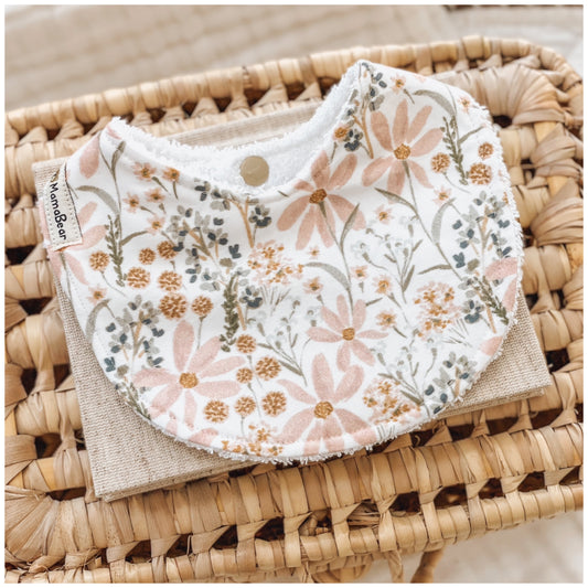 Baby girl bib featuring a delicate pastel floral print with soft peach daisies and green leaves on a white background. Displayed on a woven basket with a neutral linen cloth — a handmade bib perfect for newborn girls.
