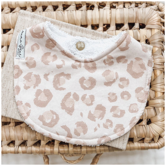 blush pink toned baby bib with pastel pink leopard print spots, laid flat on a  linen cloth and woven basket.