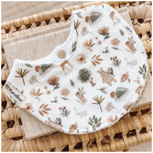 Baby bib with a woodland animal print, showing soft illustrations of bears, ducks, trees, and flowers in warm earthy colours. Displayed on a woven basket with a linen cloth — a nature-inspired handmade bib for newborns.