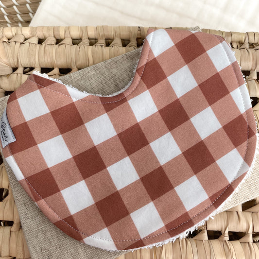 Baby bib with a rust and white gingham check pattern. Displayed on a woven basket with a neutral fabric base — a handmade, unisex baby bib ideal for newborns or gifts.