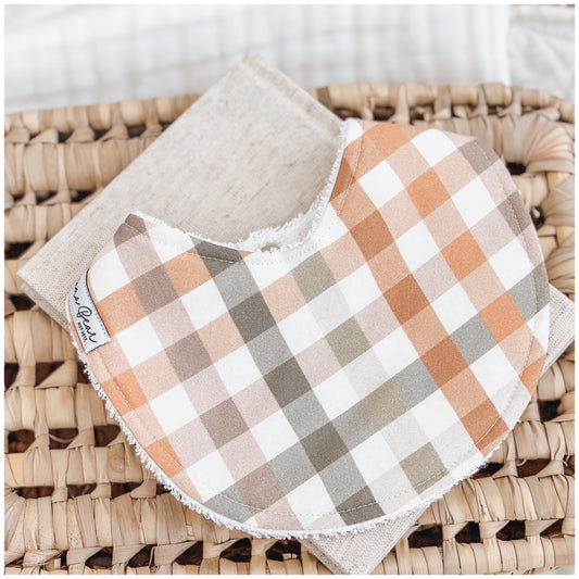 Handmade baby bib with an autumn-toned gingham print featuring rust, sage, peach, and cream checks. Bib is backed with beige terry towelling and styled on a woven basket.