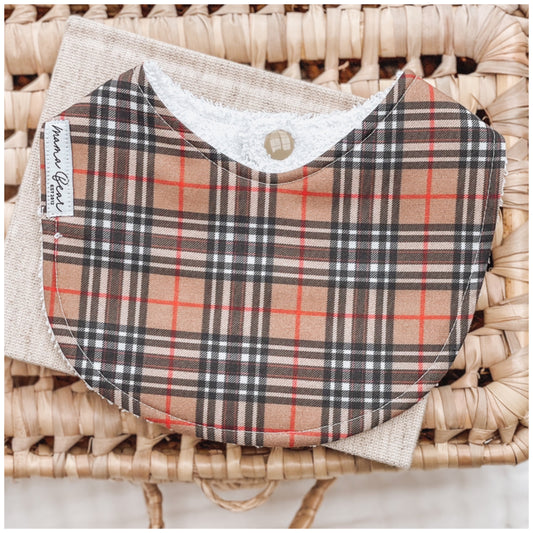 Neutral-toned baby bib with a tan, black, white and red plaid check pattern, styled on a natural linen cloth and woven basket.
