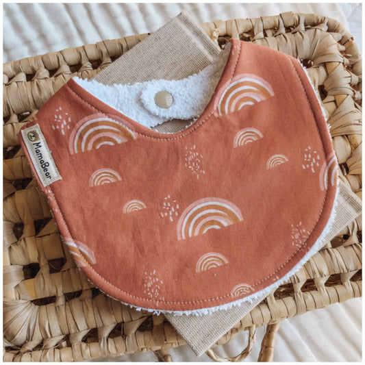 Unisex baby bib in rust orange with soft pink rainbow print. Photographed on a woven basket with a natural linen cloth — a handmade gender-neutral bib suitable for newborns and baby gifts.