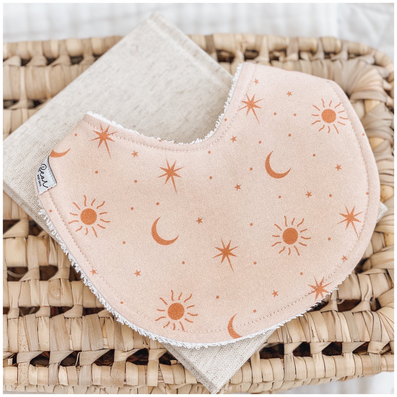 Handmade baby bib with a soft peach background and scattered burnt orange sun, moon, and star illustrations. Bib is backed with beige terry towelling and styled on a woven basket.