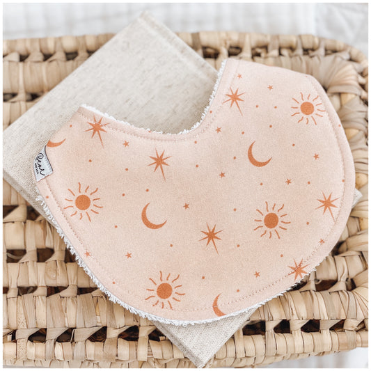 Handmade baby bib with a soft peach background and scattered burnt orange sun, moon, and star illustrations. Bib is backed with beige terry towelling and styled on a woven basket.