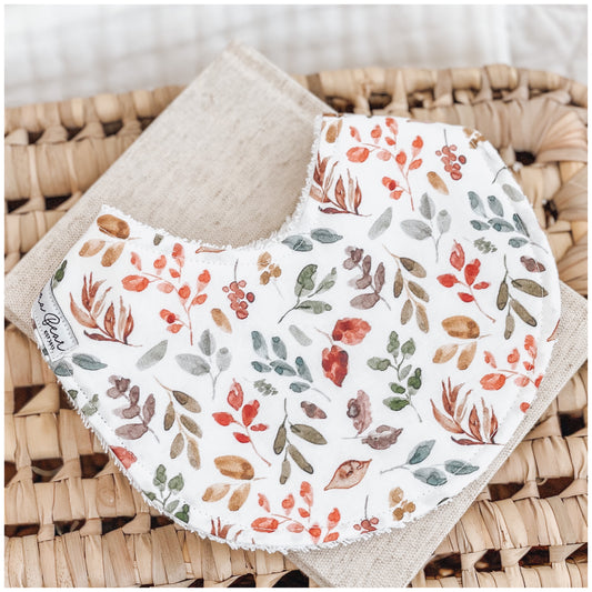 Baby bib with a scattered print of autumn leaves in soft shades of rust, green, ochre, and brown on a cream base. The bib is displayed over a natural linen cloth and wicker basket.