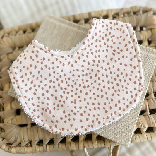 Neutral baby bib with terracotta fawn-like spots on a beige background. Handmade with cotton and terry towelling backing, displayed flatlay on woven mat.