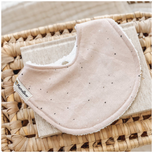 Baby bib in a soft latte colour with a scattered black speckle print. Photographed on a woven basket with a natural linen cloth — a neutral, handmade option for newborns.