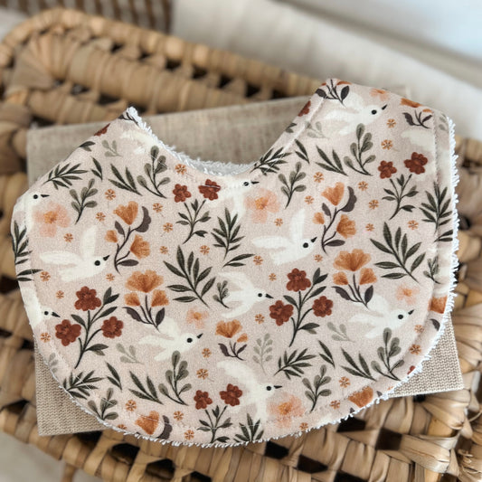 Flatlay of a handmade baby bib with a bird and floral print in earthy tones. The design features small white birds, warm-toned flowers in peach and rust, and green leafy branches on a beige background. The bib is cotton with an absorbent terry towelling backing.