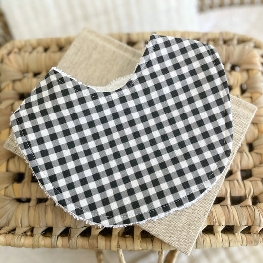 Flatlay of a handmade baby bib with a black and white gingham print. The classic check pattern gives a timeless and neutral look. Bib is made from cotton and backed with white terry towelling for absorbency.