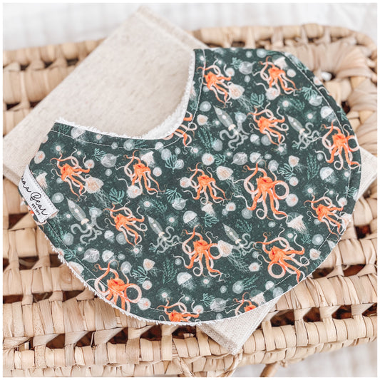 Handmade baby bib with a playful ocean-themed print showing orange octopuses, jellyfish, bubbles, and seaweed on a dark green base. Bib is backed with soft beige terry towelling and placed on a woven basket.