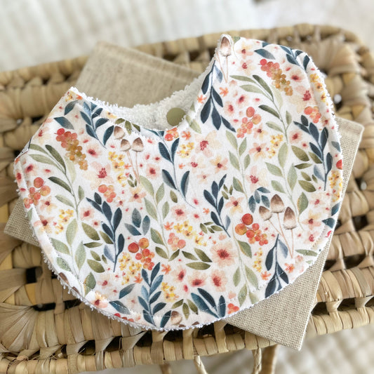 Flatlay of a handmade baby bib with a floral print. The fabric shows colourful wildflowers and green leaves in shades of peach, coral, yellow, and blue on a soft cream background. The bib is made from cotton with a white terry towelling backing.