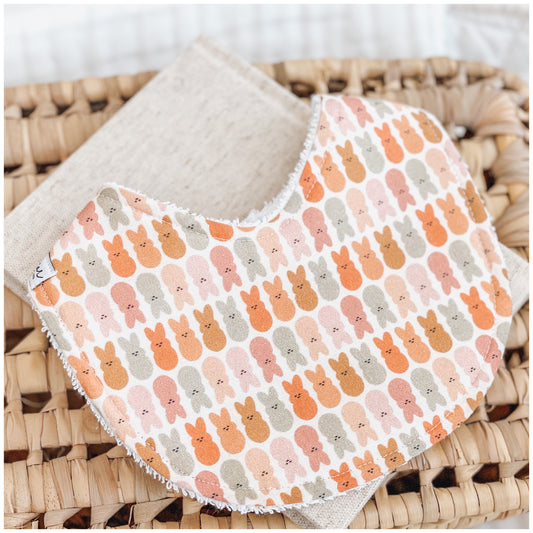 Handmade baby bib with a repeating bunny face print in warm neutral tones including peach, sage, brown, and blush on a white background. Bib is backed with beige terry towelling and shown on a woven basket.