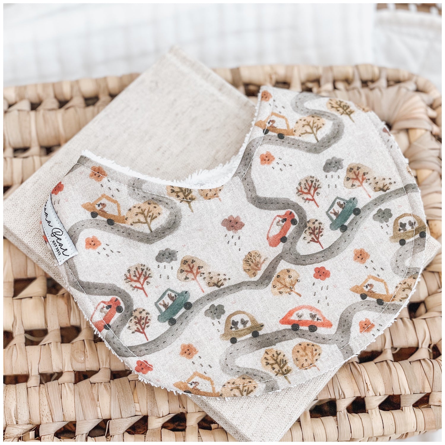 Handmade baby bib with a road trip pattern showing cars, winding roads, trees, clouds, and houses in muted rust, olive, and mustard tones on a light beige background. Bib is backed with beige terry towelling and placed on a woven basket.