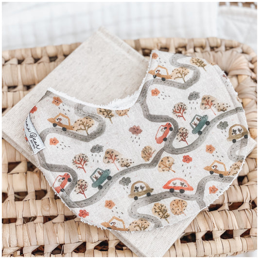 Handmade baby bib with a road trip pattern showing cars, winding roads, trees, clouds, and houses in muted rust, olive, and mustard tones on a light beige background. Bib is backed with beige terry towelling and placed on a woven basket.