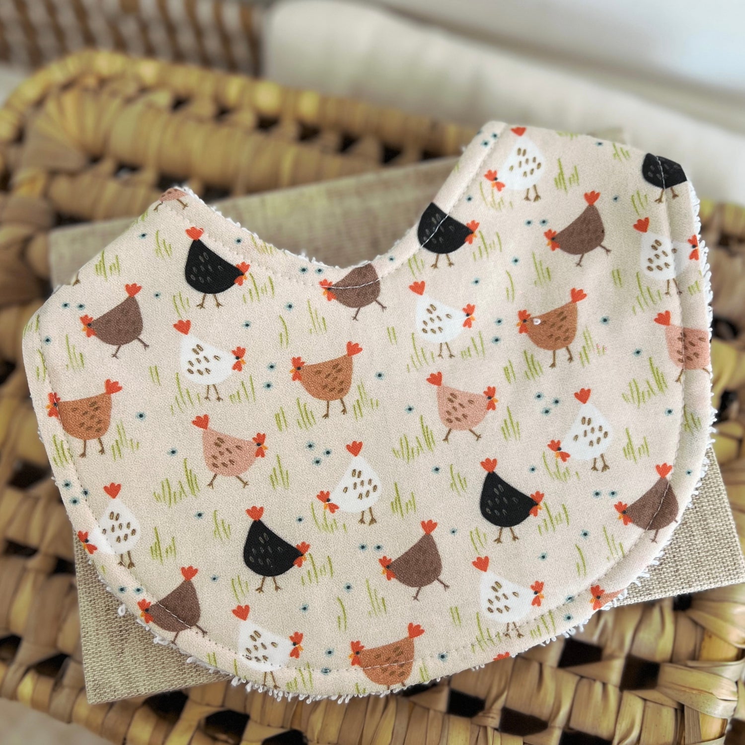 Flatlay of a handmade baby bib with a chicken-themed print. The design features cartoon-style chooks in shades of white, brown, tan, and black scattered across a light beige background with grassy green details. Made from cotton with a terry towelling backing for absorbency.