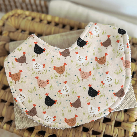 Flatlay of a handmade baby bib with a chicken-themed print. The design features cartoon-style chooks in shades of white, brown, tan, and black scattered across a light beige background with grassy green details. Made from cotton with a terry towelling backing for absorbency.