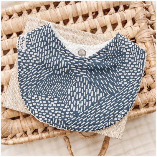 Dark blue baby bib featuring a scattered dash and star-like geometric pattern in white, styled on a neutral-toned fabric and wicker basket.