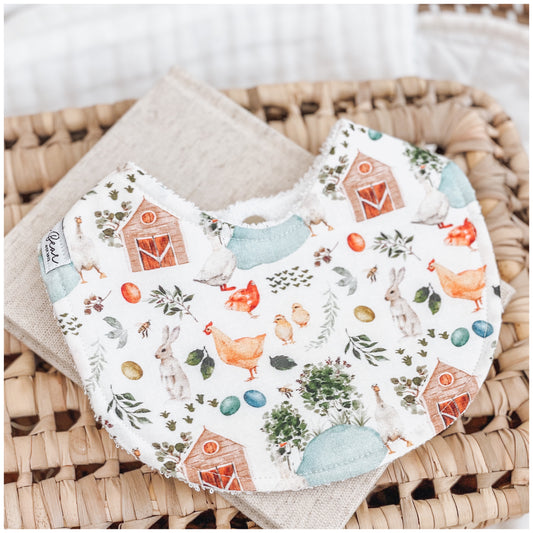 Handmade baby bib with a farm-themed print including chickens, chicks, bunnies, barns, watering cans, plants, and pastel-coloured eggs on a white background. 