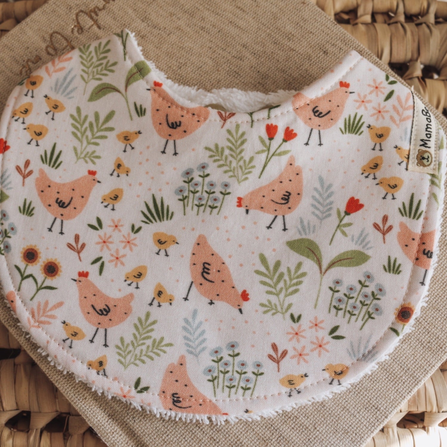 Handmade baby bib with a soft peach and cream base, printed with chickens, baby chicks, and delicate greenery, styled on natural textured fabric.