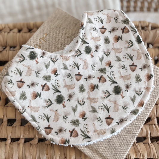 Neutral-toned baby bib with a woodland pattern of rabbits, acorns, mushrooms, leaves, and greenery on a cream background. Backed with terry towelling, ideal for dribbles, teething, and newborn feeding.