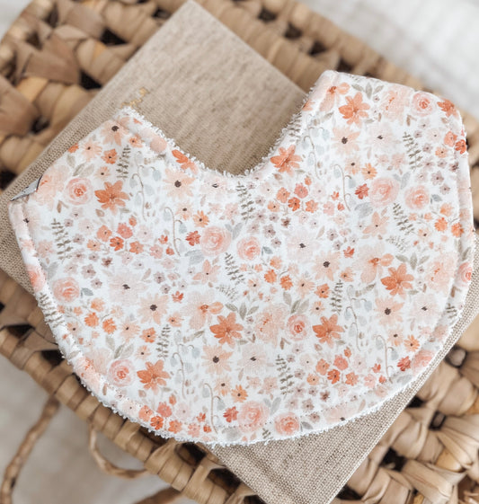 Baby bib with a soft floral pattern including blush and peach flowers, muted greenery, and small bluebell details on a cream background. Terry towelling backing for absorbency—ideal for newborns and teething.
