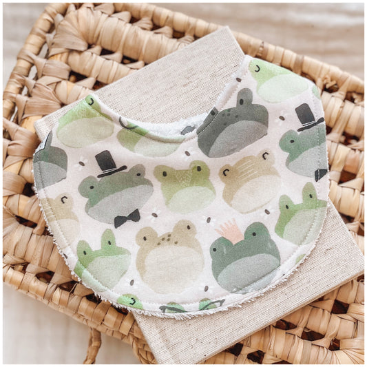Neutral baby bib with a cute frog print, including green and beige frogs wearing top hats, crowns, and bowties, designed for babies and toddlers. Set on a soft white background.
