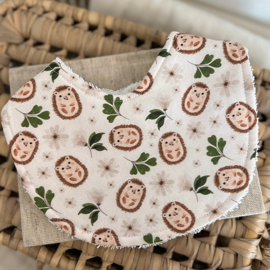 Flatlay of a handmade baby bib with a whimsical hedgehog pattern. The fabric features smiling brown hedgehogs, dainty beige flowers, and green leafy sprigs on a light cream base. Bib is cotton with a soft terry towelling reverse side for absorbency.