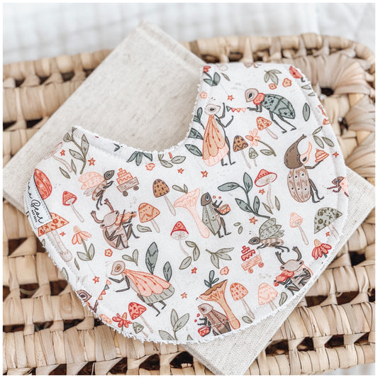 Handmade baby bib with whimsical bug illustrations, including insects pushing cakes, mushrooms, leaves, and woodland-themed doodles in warm earthy tones on a soft cream base. Displayed on a beige cloth over a woven basket.
