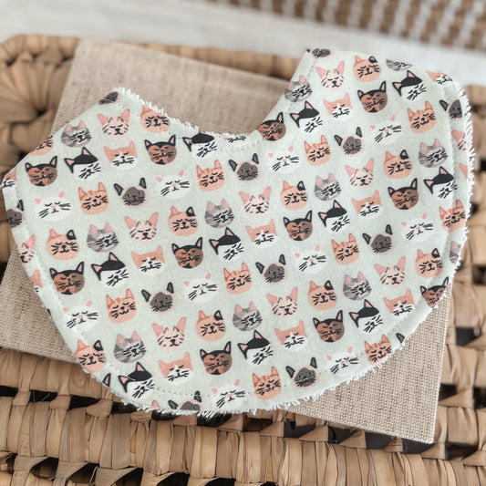 Flatlay of a handmade baby bib with a kitten face print. The design shows rows of illustrated cat faces in blush, grey, black, and peach shades on a light mint or soft sage background. Bib is backed with white terry towelling for added absorbency.