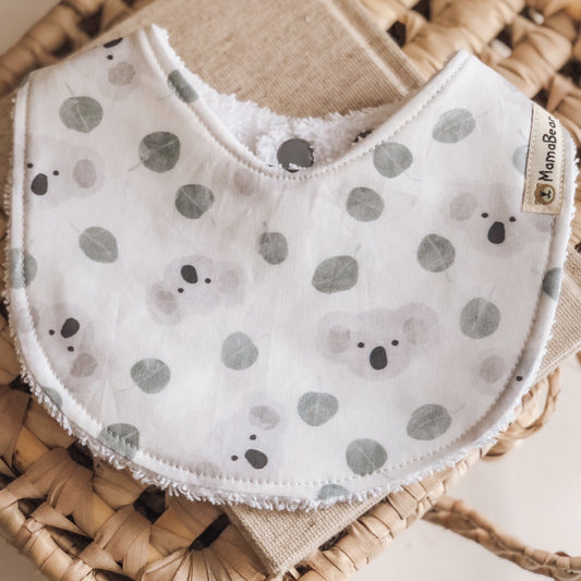 Handmade baby bib with soft grey koalas and green eucalyptus leaves on a white cotton background, backed with white terry towelling.