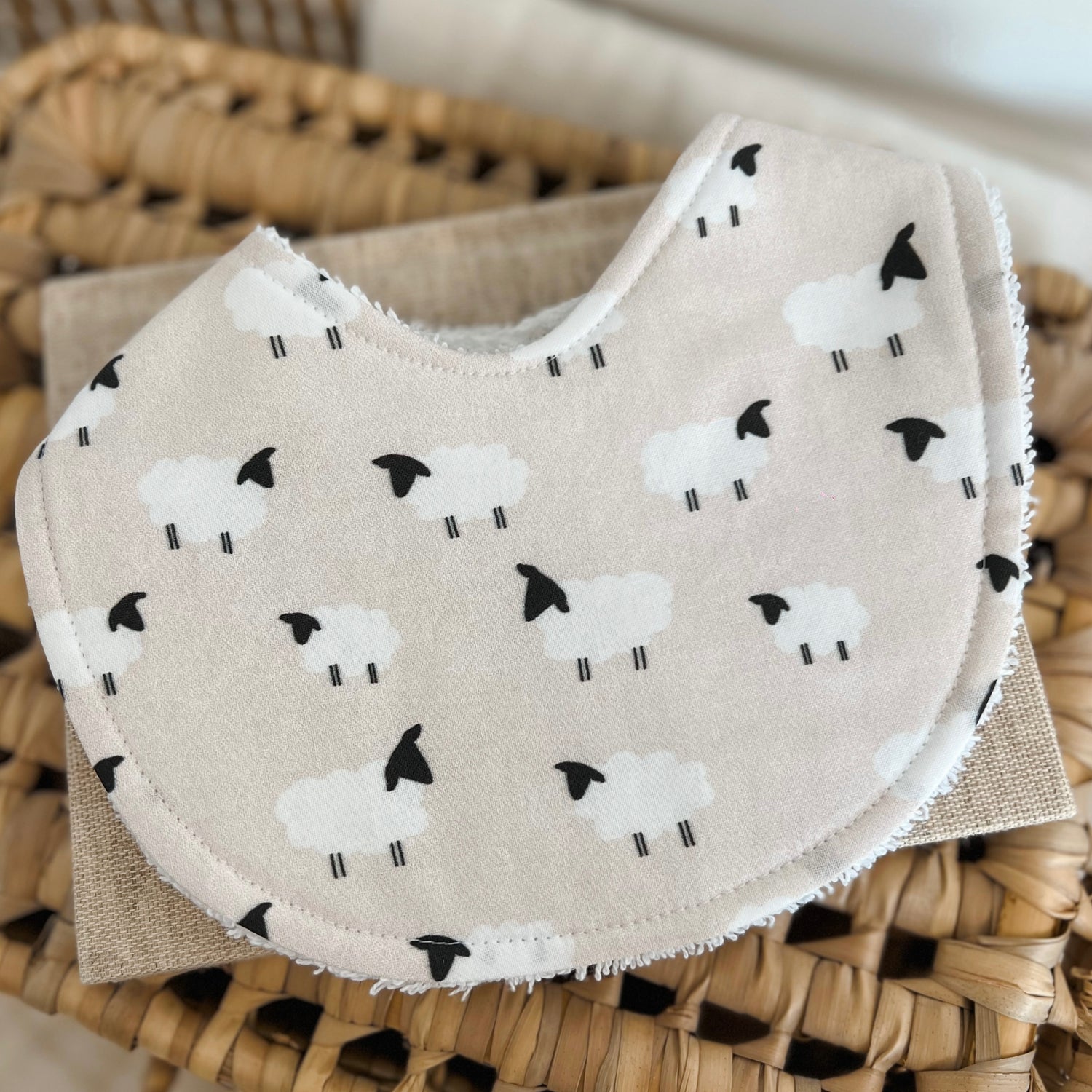 Handmade baby bib featuring a simple sheep print for farm lovers. The fabric shows fluffy white lambs with black faces and legs, evenly spaced on a beige background. Bib has a cotton front and terry towelling backing for absorbency.