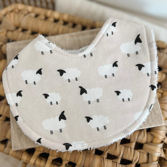Handmade baby bib featuring a simple sheep print for farm lovers. The fabric shows fluffy white lambs with black faces and legs, evenly spaced on a beige background. Bib has a cotton front and terry towelling backing for absorbency.