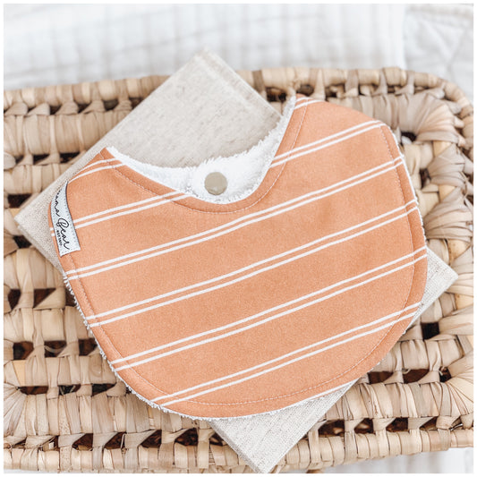 Rust-coloured baby bib with white diagonal stripes, laid flat on a neutral linen cloth inside a woven basket.