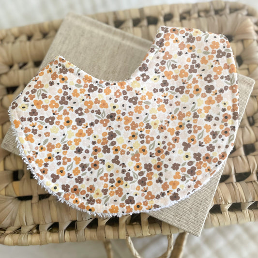 Handmade baby bib with tiny scattered flowers in brown, mustard yellow, and burnt orange on a soft cream background. Laid flat on a neutral fabric and woven basket.