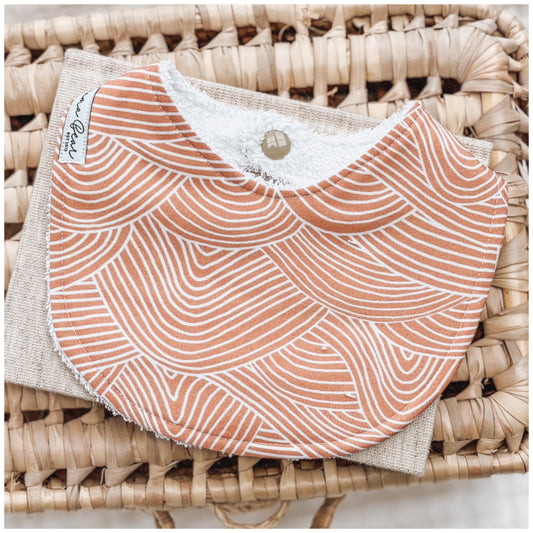 Rust orange baby bib with white curved wave-like lines in an abstract pattern, displayed on neutral-toned props in a woven basket.