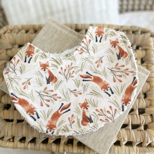 Flatlay of a handmade baby bib featuring orange foxes surrounded by leaves and grass on a cream background. The foxes are illustrated in different poses across the fabric. Bib is cotton with a white terry towelling backing for absorbency.