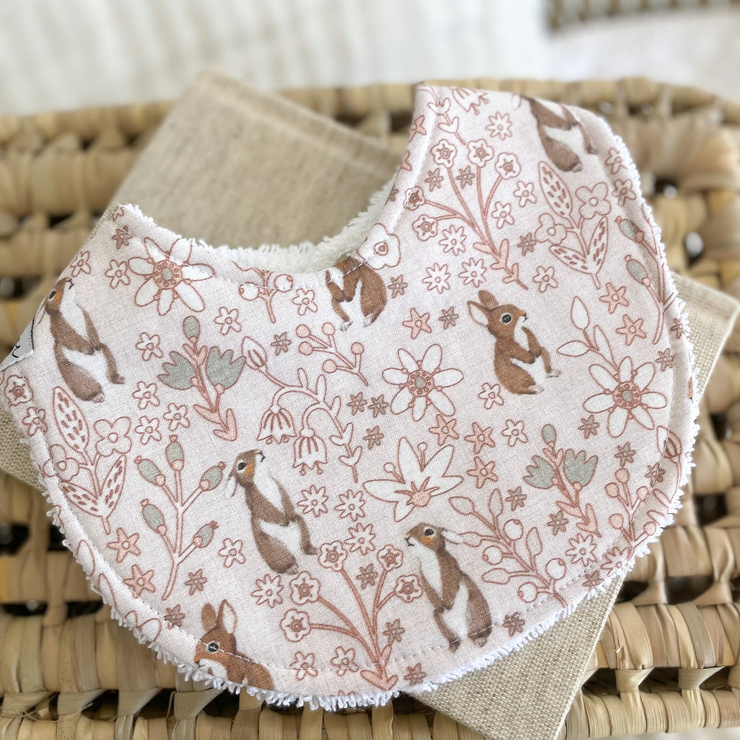 Flatlay of a handmade baby bib with a light pink background, covered in dainty floral line drawings and brown bunny rabbits. The bib is made from soft cotton with a white terry towelling back for absorbency.