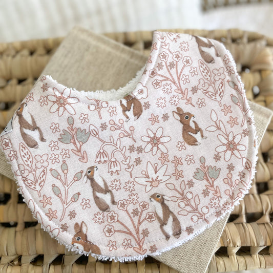 Flatlay of a handmade baby bib with a light pink background, covered in dainty floral line drawings and brown bunny rabbits. The bib is made from soft cotton with a white terry towelling back for absorbency.