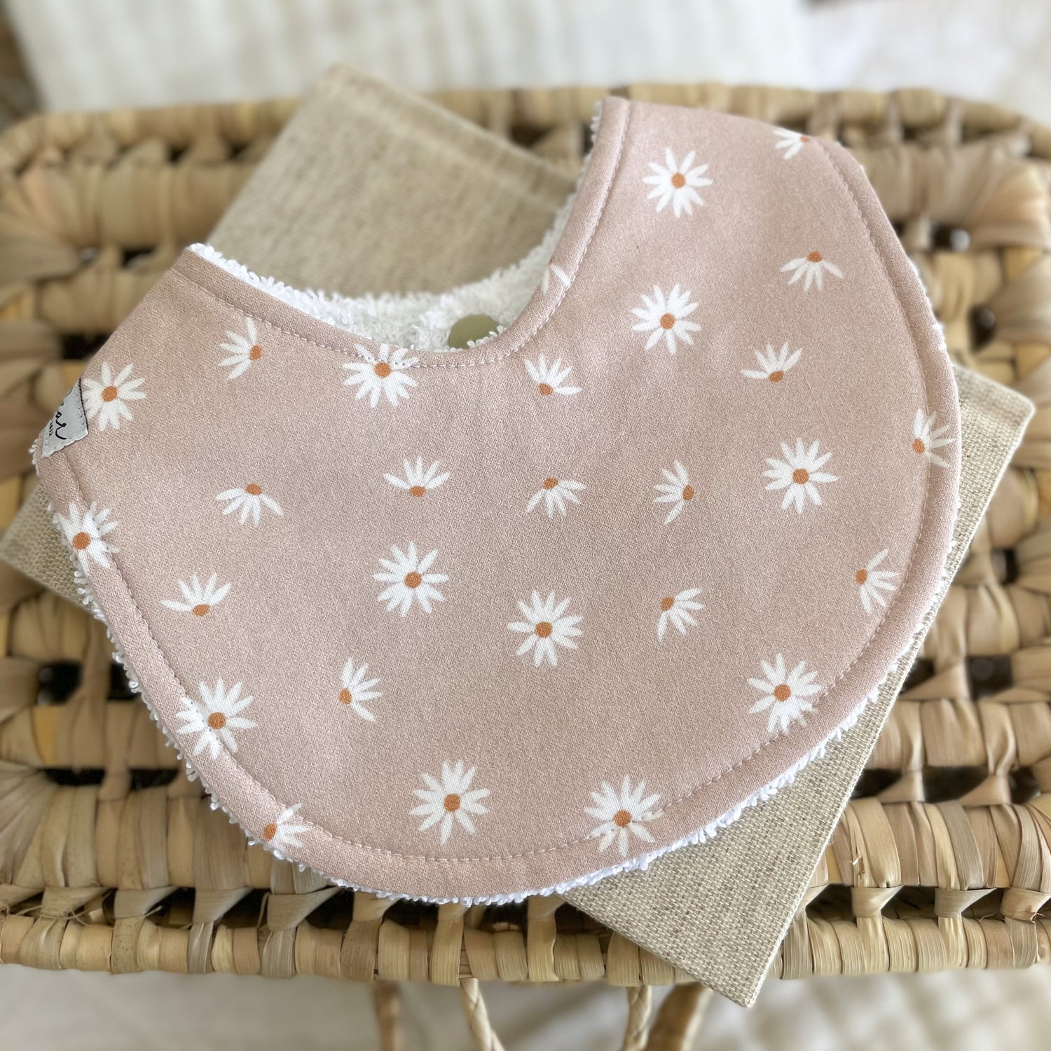 Flatlay of a handmade baby bib with a blush pink background and simple white daisy print. Each daisy has a warm brown centre. The bib is made from soft cotton with a white terry towelling back for absorbency.