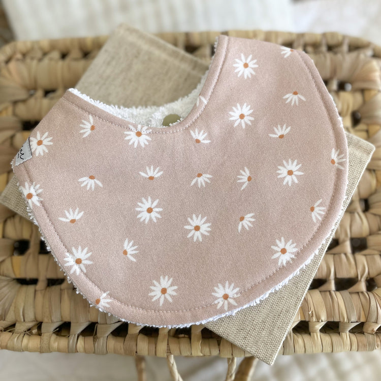 Flatlay of a handmade baby bib with a blush pink background and simple white daisy print. Each daisy has a warm brown centre. The bib is made from soft cotton with a white terry towelling back for absorbency.