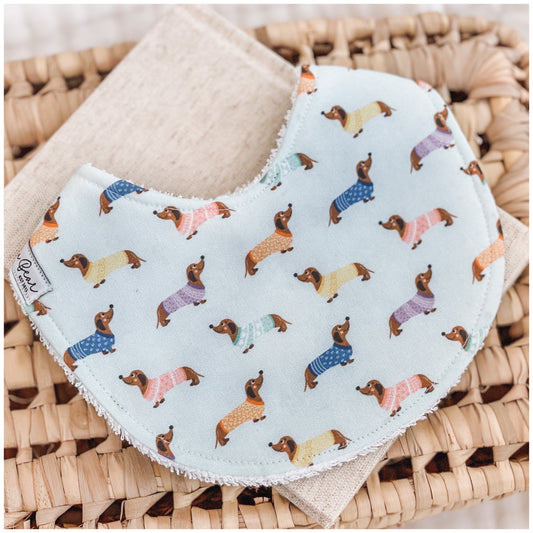Handmade baby bib with a pale blue background and a repeating dachshund print. The dogs wear jumpers in pastel colours including pink, yellow, blue, and lavender. Bib is backed with beige terry towelling and styled on a woven basket