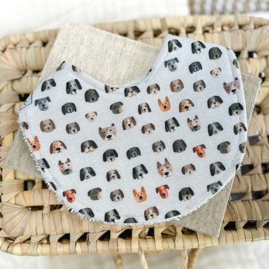 Flatlay of a handmade baby bib with a dog-themed print. The design includes cartoon-style puppy faces in shades of tan, grey, black, and rust on a white background. Bib is made from cotton and backed with absorbent terry towelling.