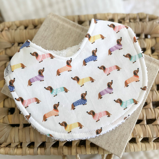 Flatlay of a handmade baby bib featuring dachshund dogs in colourful jumpers on a white background. Each sausage dog is illustrated in a rainbow-coloured sweater. The bib is made from cotton and backed with white terry towelling for absorbency.
