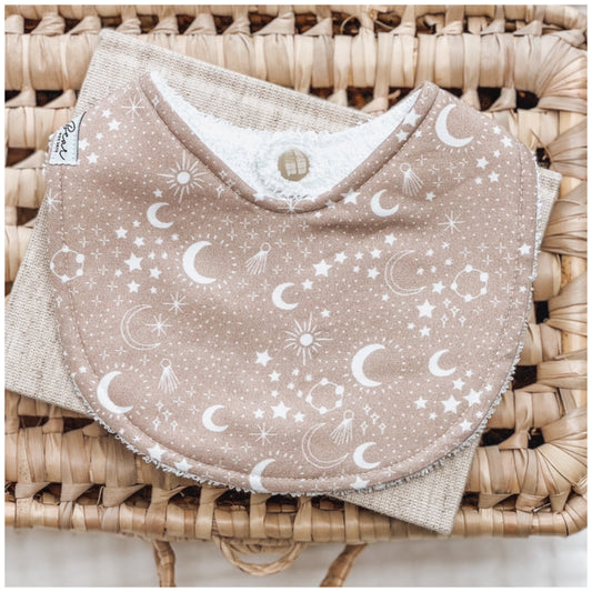 Neutral beige baby bib with white celestial pattern of moons, stars, constellations, and shooting comets, photographed on a natural woven basket.