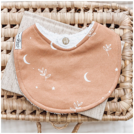 Rust brown / ochre baby bib with white crescent moons, stars, and simple leafy branch illustrations, displayed on natural linen over a wicker basket.