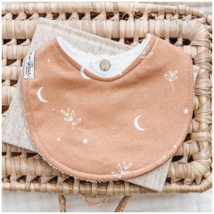 Rust brown / ochre baby bib with white crescent moons, stars, and simple leafy branch illustrations, displayed on natural linen over a wicker basket.