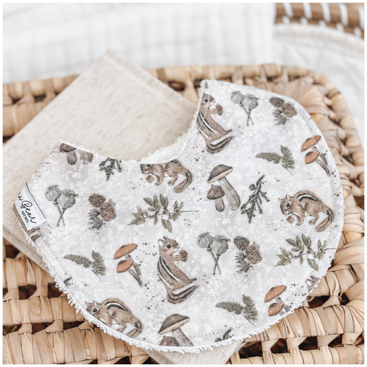 Handmade baby bib with a forest-themed print showing chipmunks, mushrooms, and leafy foliage in neutral tones on a soft mottled beige background. Bib is backed with beige terry towelling and styled on a woven basket.