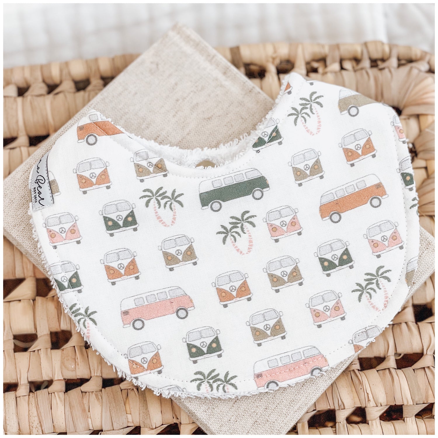 Unisex baby bib with a print of retro vans and palm trees in muted pastel tones on a soft white background, laid flat on a woven basket.